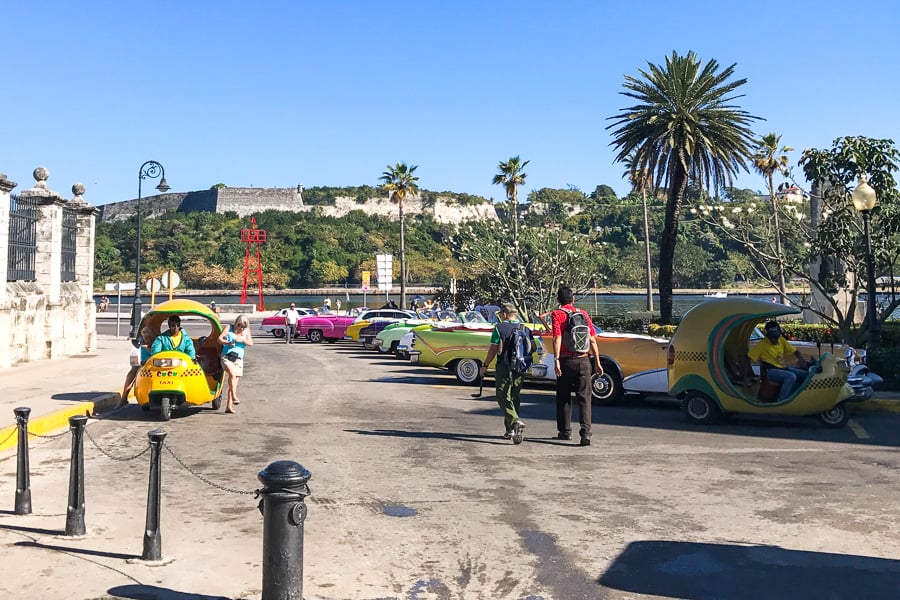 A traveller visiting Cuba gets out of a yellow coco taxis opposite a row of colourful classic cars. 