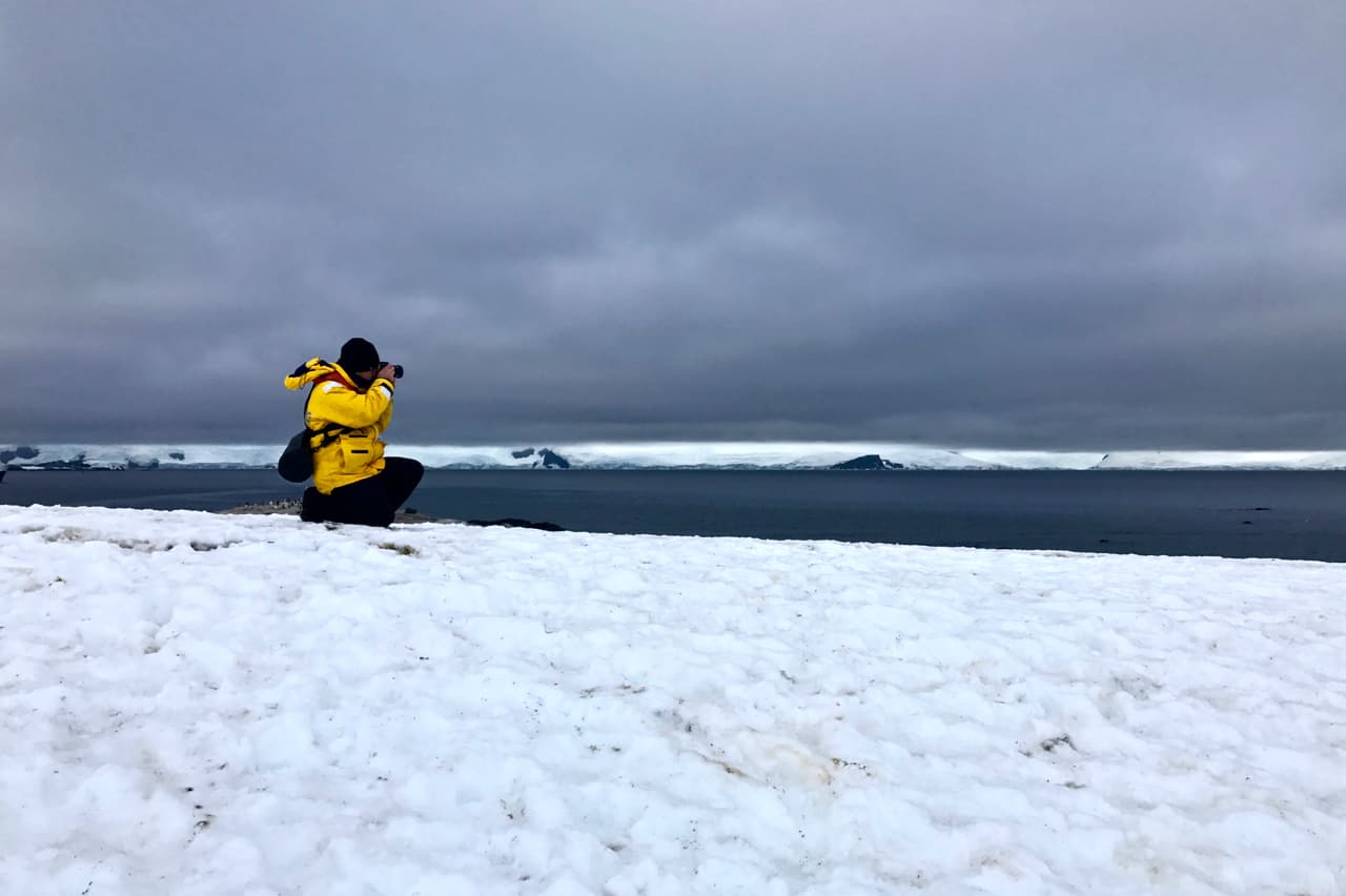 A photographer in a yellow parka kneels on the snow to capture a scene in Antarctica.