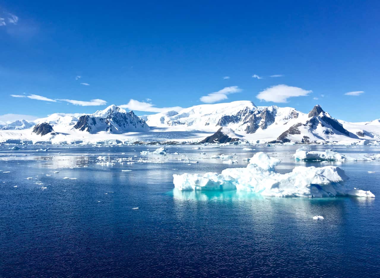 An expansive Antarctic panorama of deep blue sea and white snow-capped mountains with an iceberg in the foreground.