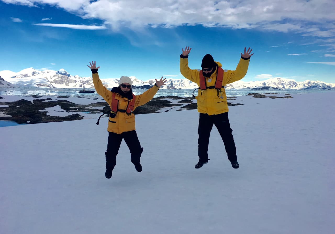 Couple dressed in yellow parkas jumping on the snow while visiting Antarctica.