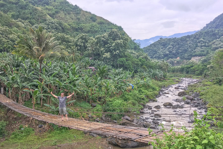 A visitor in a lush valley crossing a bamboo bridge over fast running brown water surrounded by hills on our Flores trip.