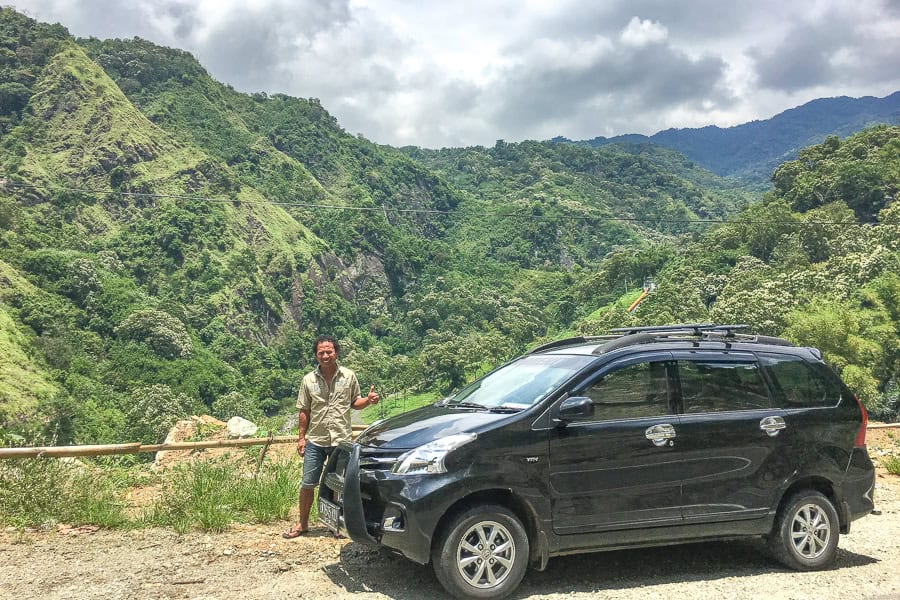 A driver stands beside a black car with a lush mountain backdrop – consider a driver for your Flores Indonesia itinerary.