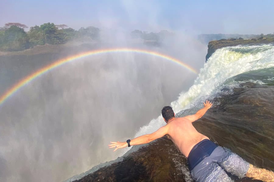 A person lays on the edge of a waterfall with their arms out with a rainbow and mist - one of the most thrilling adventures of the world.