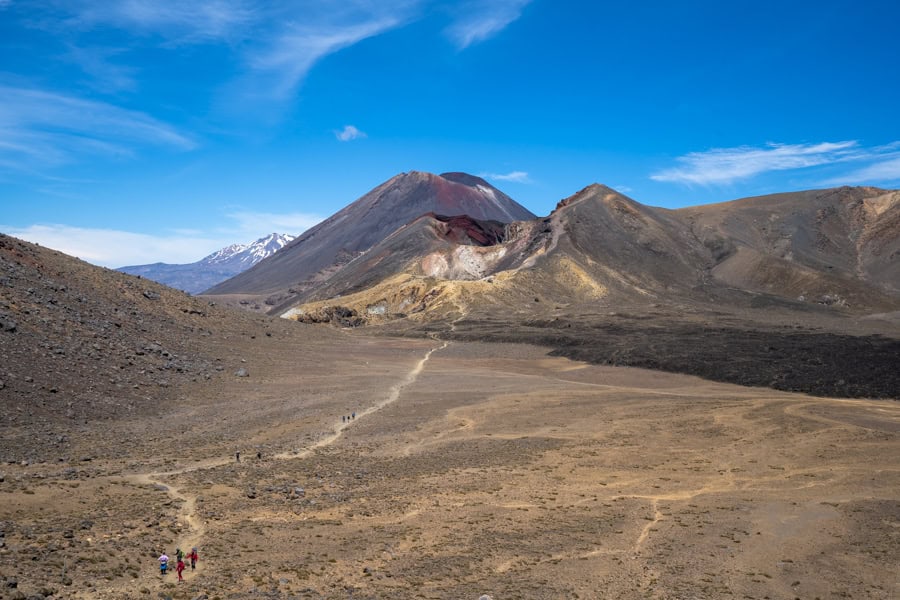A walking trail with groups of people winds in front of two cone volcanoes in New Zealand, one of the world's great adventure destinations.