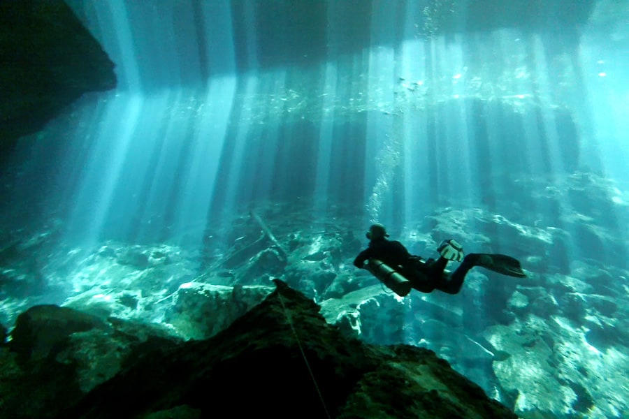 A diver is silhouetted as blue beams of light cut through the water of a cenote.