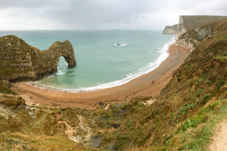 A beach with a large rocky arch stretching out into the water and high white cliffs to the right.