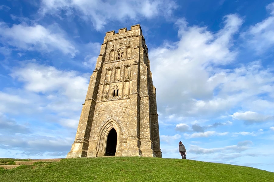 A stone tower sits atop Glastonbury Tor with a person standing next to it. 
