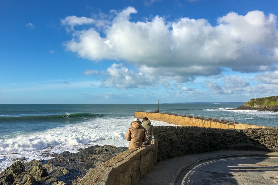 People lean against a wall watching surfers at Porthleven on our England road trip itinerary.