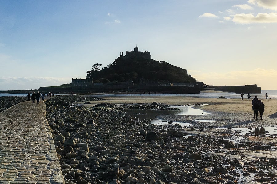 People walking across a cobbled path and rocky beach with St Michael's Mount in the background.