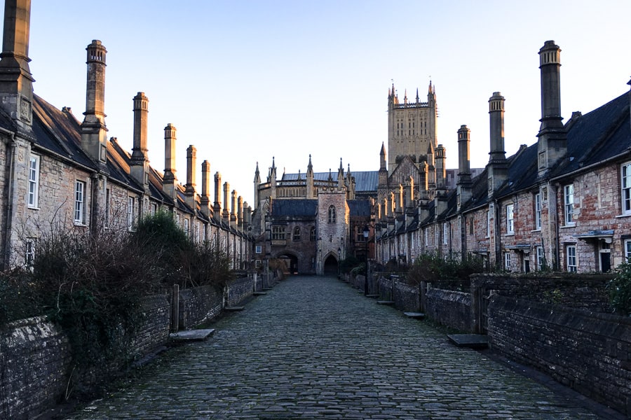 Stone cottages with chimneys line both sides of a cobbled road with Wells Cathedral in the background on an England road trip.
