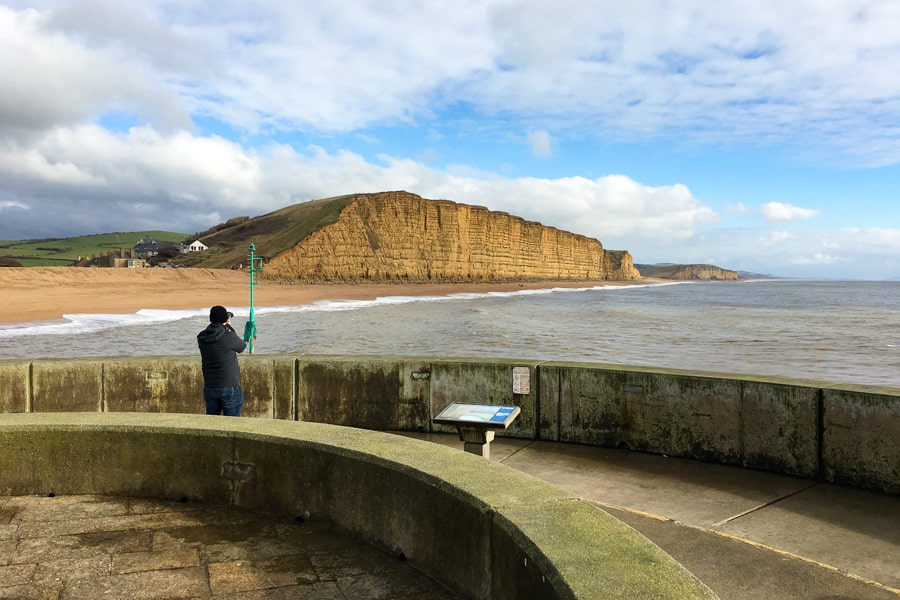 A person standing on a concrete wall looks across water to mustard-coloured cliffs of West Bay on a UK road trip itinerary.