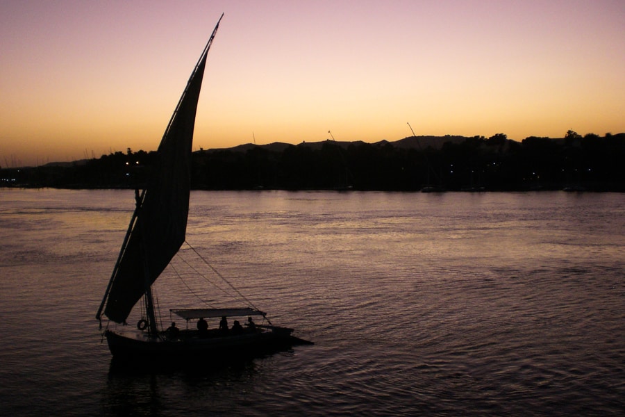 A felucca sails on the Nile beneath a pink and orange twilight sky, a must on an Egypt travel itinerary. 