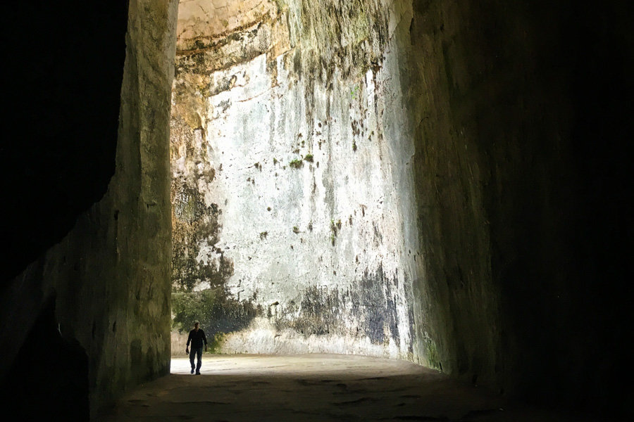 Looking out through the high, narrow entrance of the Ear of Dionysius cave as a man walks in.