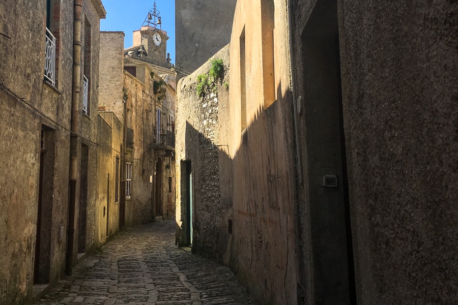 Sicily Itinerary – A narrow cobblestone street passes between the walls of houses with a clock tower rising above.  