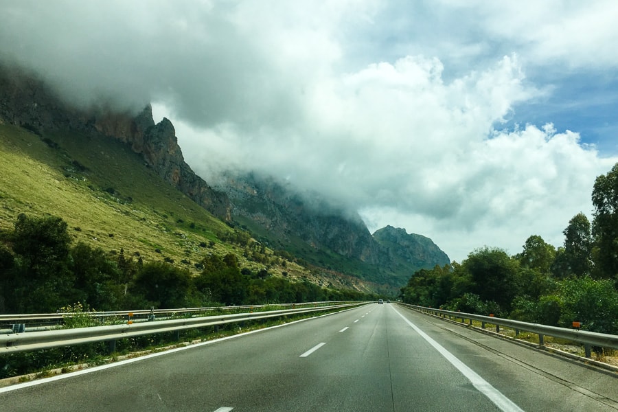 Sicily road trip – A road stretches away into the distance with cloud-capped mountains rising on the left. 