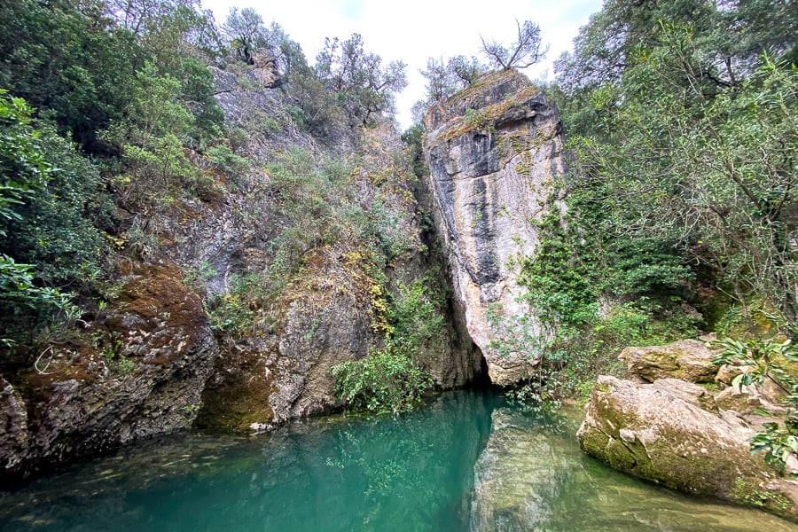 An imposing limestone wall surrounded by lush green forest is reflected in the emerald waters of Su Gologone Spring.
