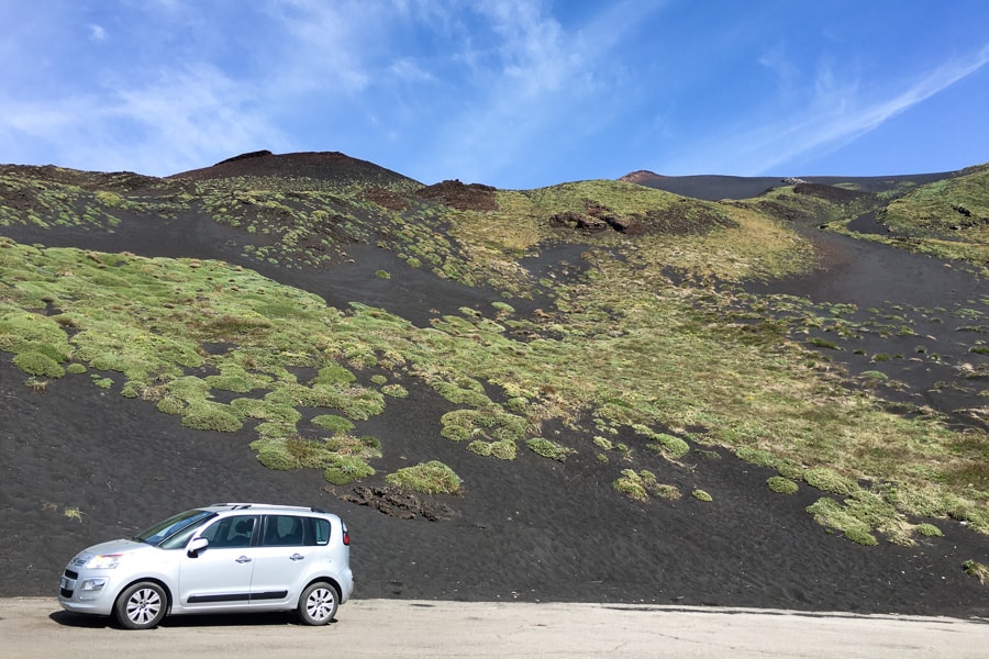 A silver car in front of a black lava field with green grass patches on Mt Etna in east Sicily. 