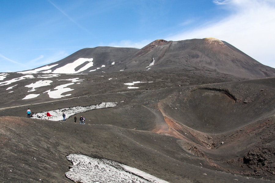 People walk along a ridge of black sand with patches of dirty snow behind them on Mount Etna. 