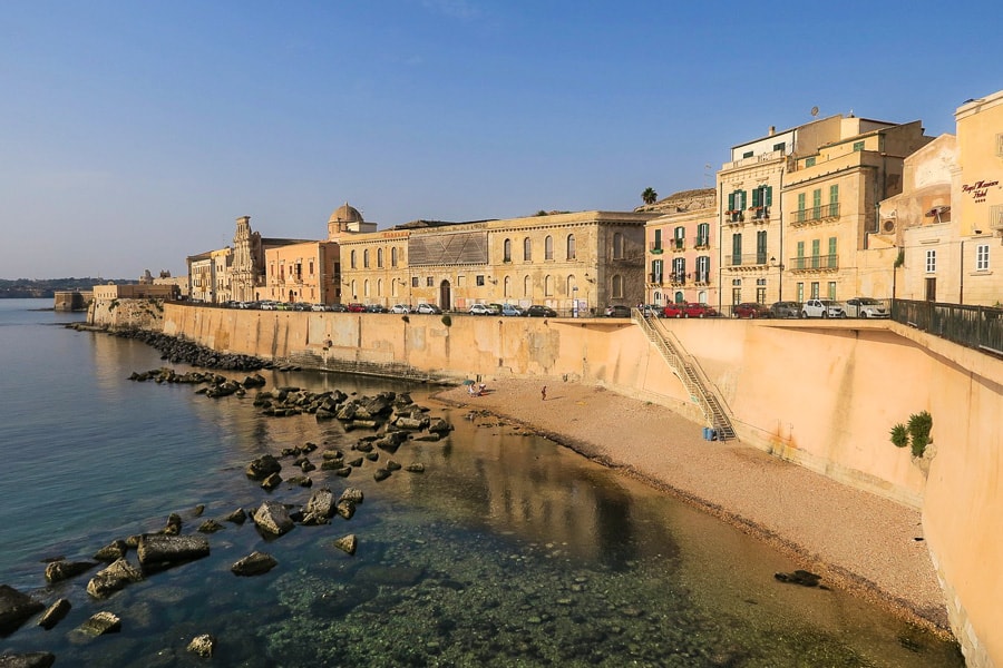 A street of old multi-level houses in Trapani stretches away above a sea wall lapped by water and a small pebbly beach. 