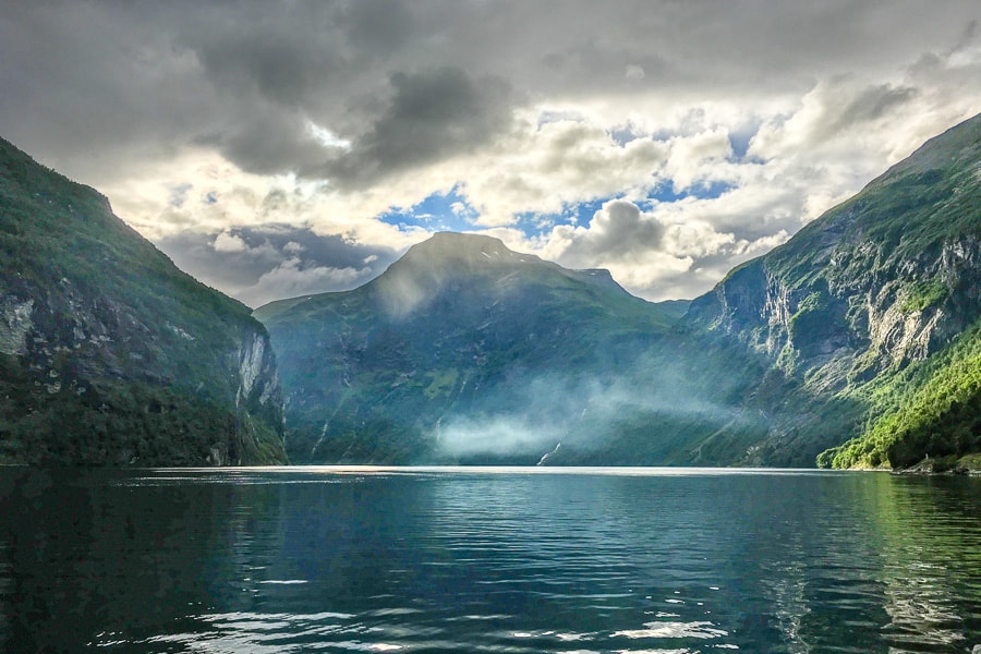 The sun breaks through the mist and clouds across Geirangerfjord with mountains in the background.