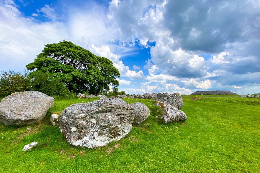 A tomb at Carrowmore sits in the foreground surrounded by grass with the large Listoghil cairn in the distance.