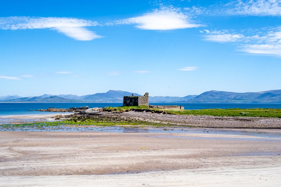 The 16th century ruins of Ballinskelligs Castle overlooking a calm bay with distant mountains on a sunny day.