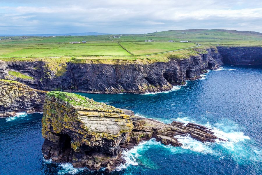 Aerial view of a rocky sea stack at Kilkee Cliffs in County Clare, Ireland with green fields in the background.