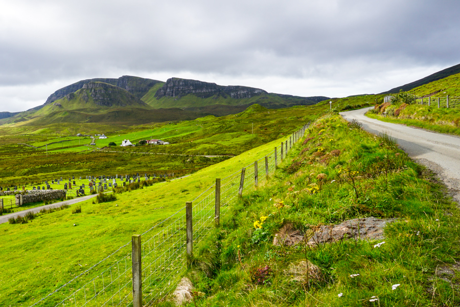 A road next to grassy fields with mountains in background on a Skye itinerary.