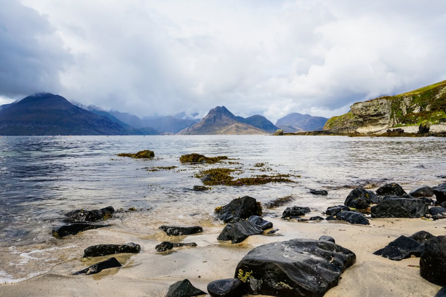A rocky beach with mountains across the water in the distance.