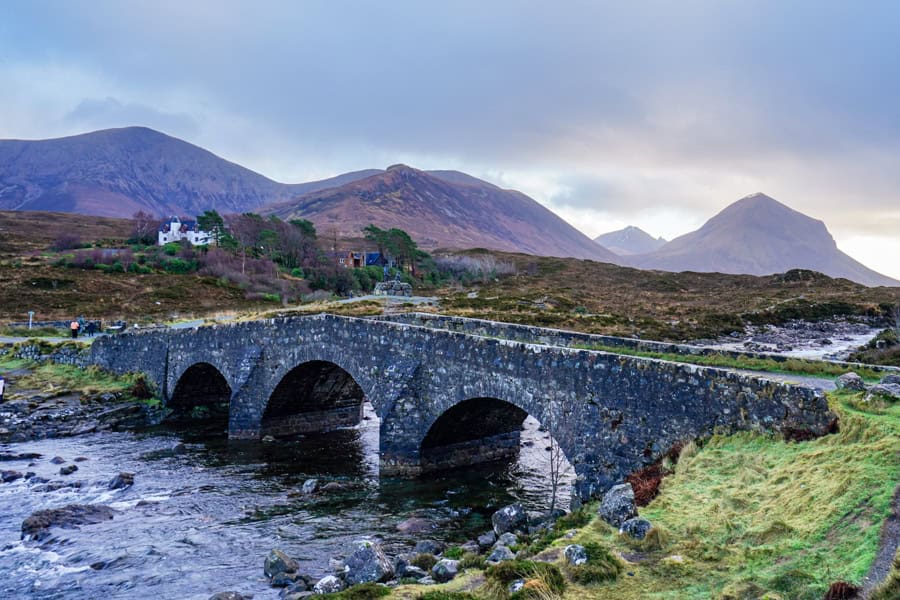 A bridge over a river and a white Skye accommodation lodge with mountains in the background.