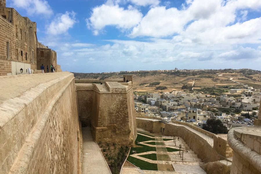 A stone fortress with a stone rampart overlooking a village and countryside in the background.