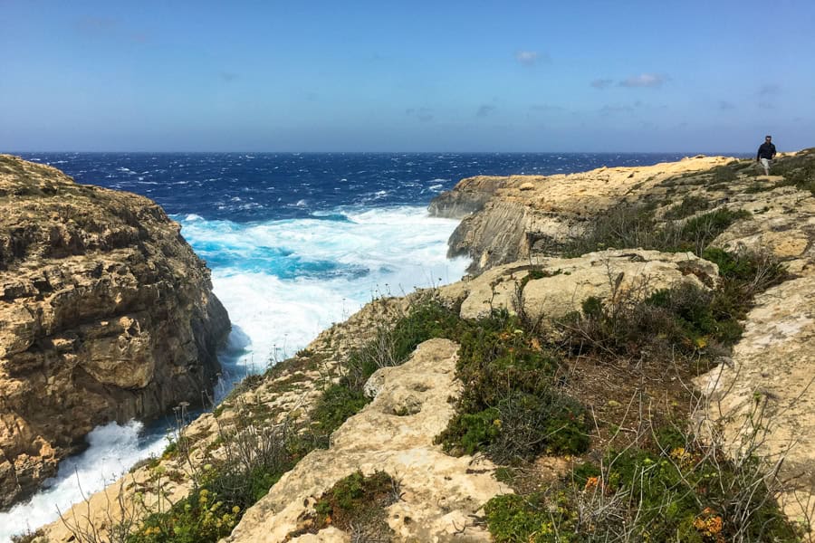A man walks along cliffs by a narrow canyon with waves crashing.