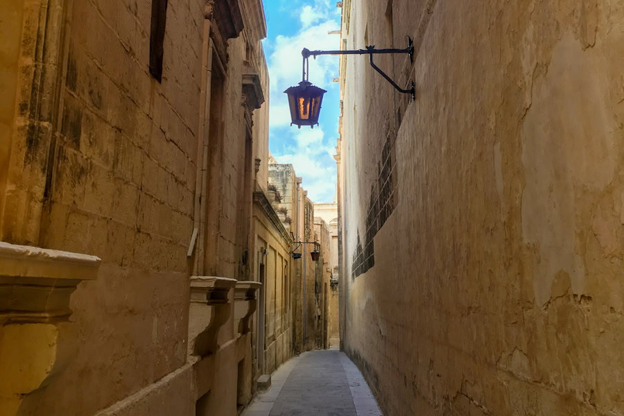 A narrow alley weaves between high walls with an overhead lamp in Mdina.
