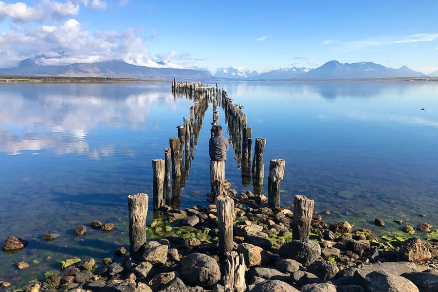 A person sits on the post of a long former pier over the water in Puerto Natales, a top Patagonia travel stop.