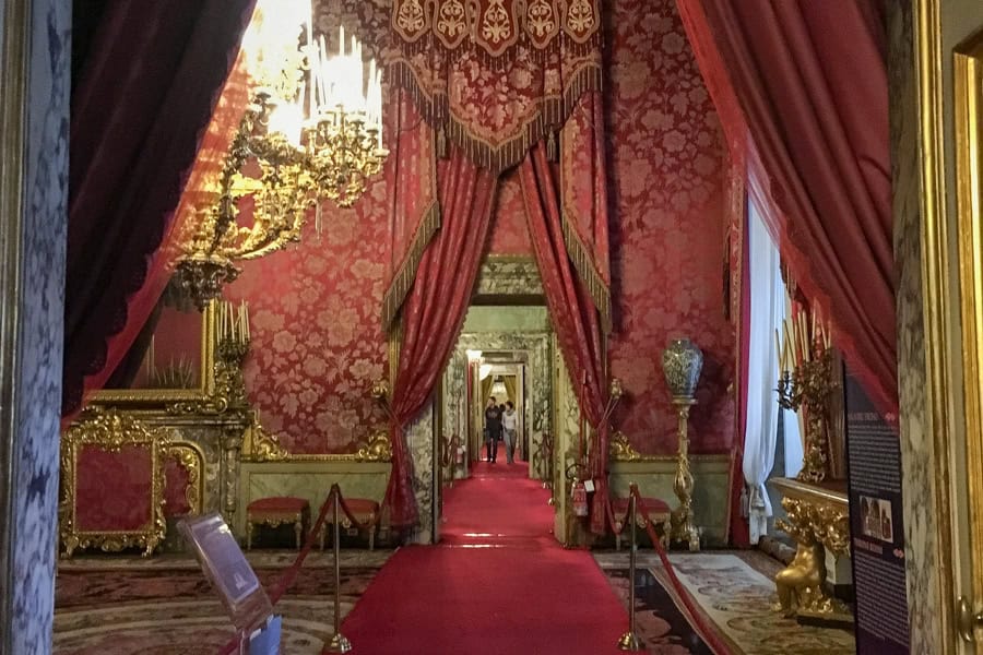 A couple visiting Florence Pitti Palace approach a room with red carpet, curtains and wall coverings.