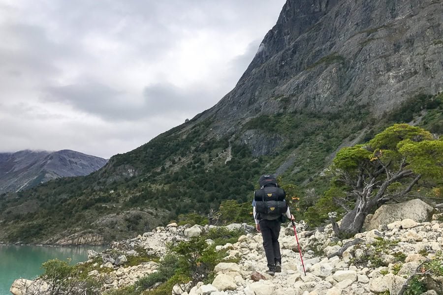 A person hikes on the rocky trail between a lake and a mountain on the W Hike Patagonia.
