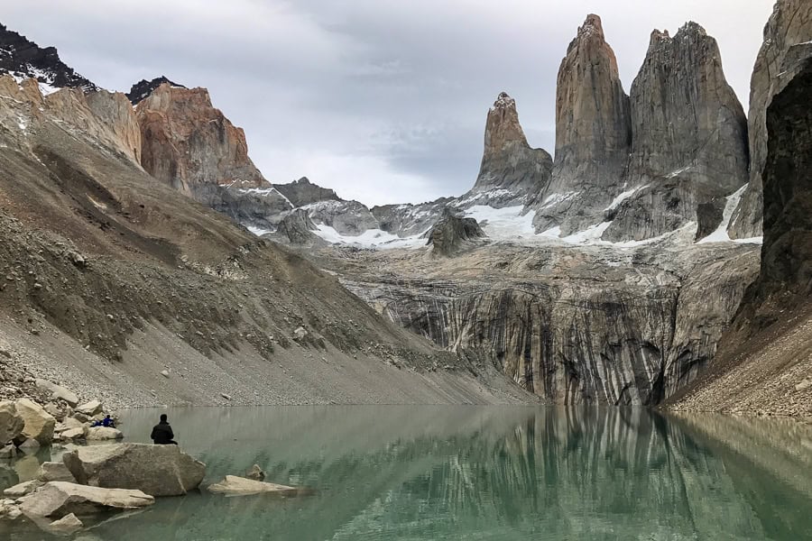 A person sits on a boulder at the edge of a lake looking at the three peaks of Las Torres.