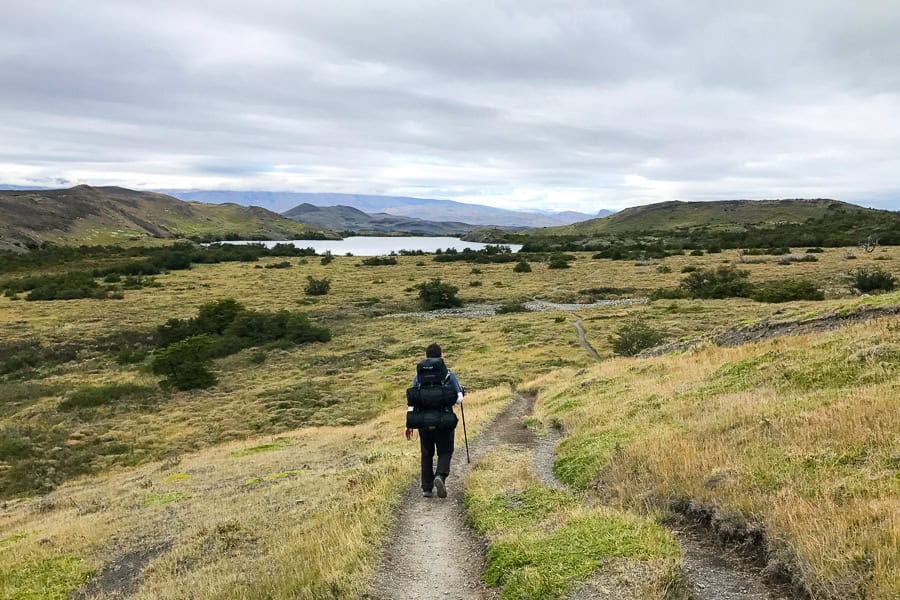 A hiker with a pack walks along a trail with grassy hills and lake views on the W trail Patagonia.