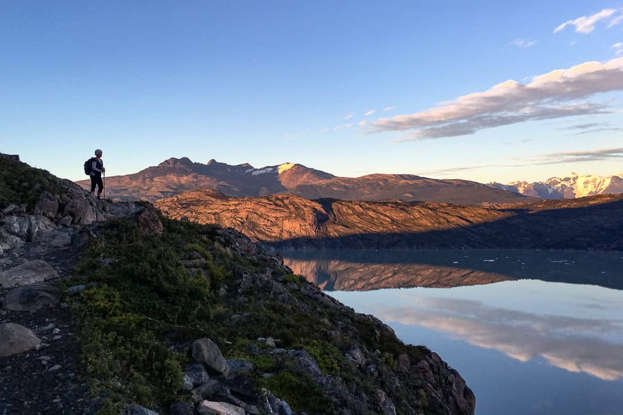 A person stands above a lake reflecting the sky in the early morning while hiking Torres del Paine.