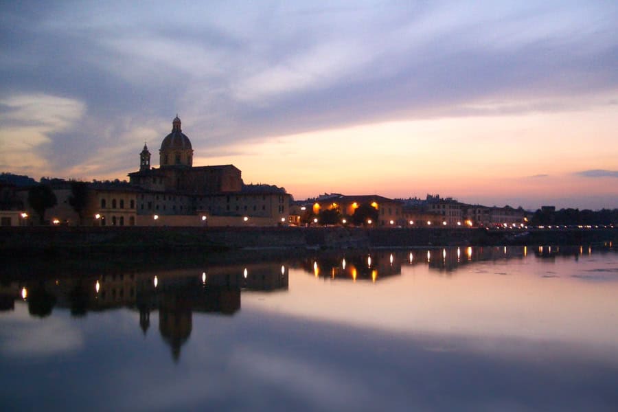 A church and buildings with lights line the River Arno in Florence at twilight.