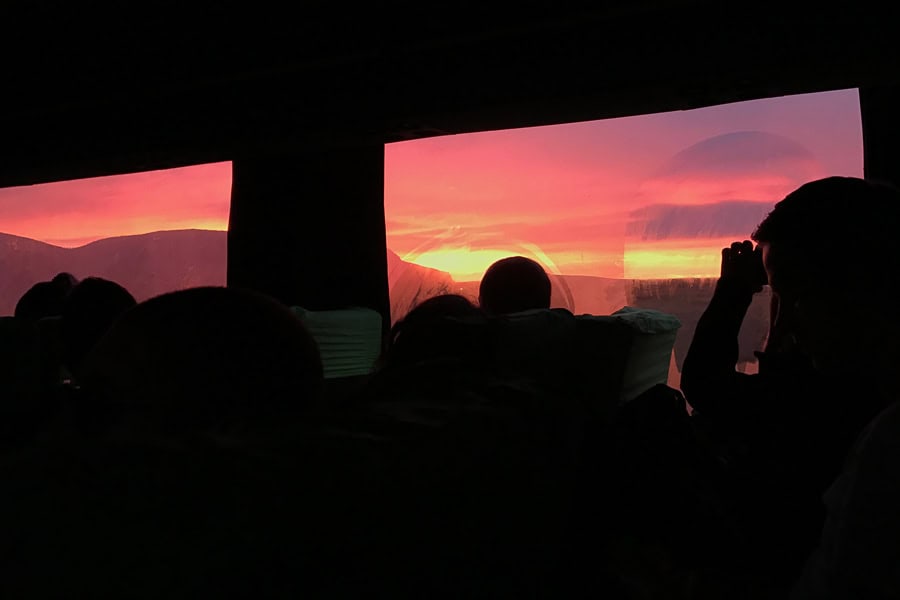 Silhouette of people looking through bus windows at a pink dawn sky.