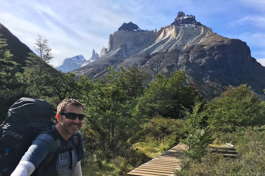 A person stands in the foreground on a wooden track with a huge mountain rising in the background on the W hike Patagonia.