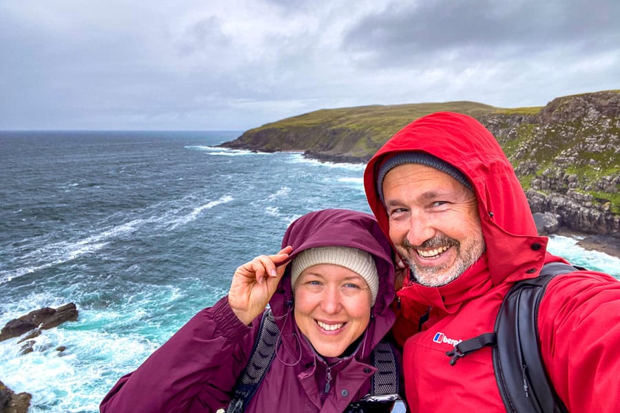 Visitors at Stoer Head on the NC500 route are buffeted by wind and rain on a cloudy day with coastal cliffs in the distance.