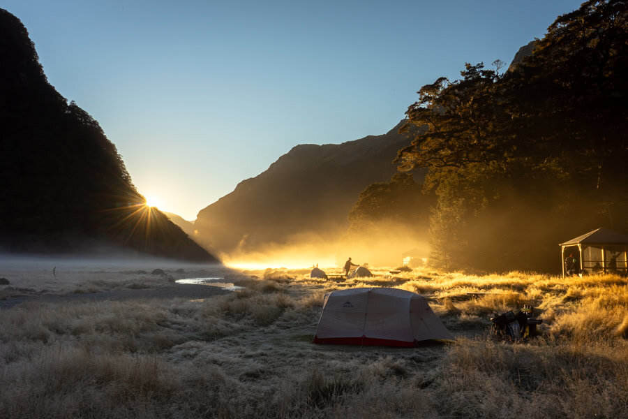The sun rises over a misty campsite with a tent and some campers at the Routeburn Flats hut and campsite area.