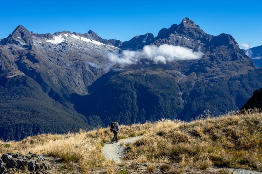 A person walking on the Routeburn Track past an orange post with mountains rising in the background.