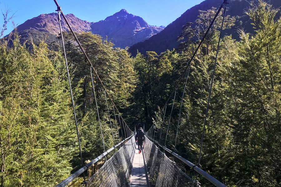 A hiker crosses a swing bridge with a mountain in the background: New Zealand Great Walks.