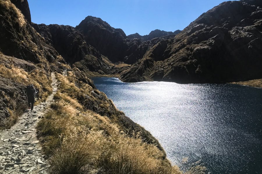 A hiker walks along a narrow rocky path next to a mountain lake hiking New Zealand.