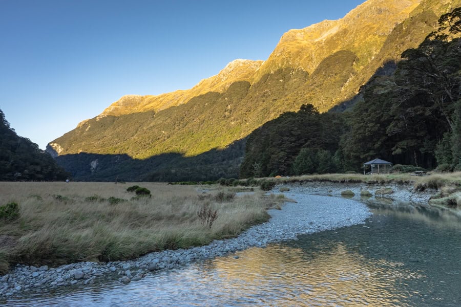 A tent and shelter sit beside a river running through a valley with mountains behind on one of the NZ Great Walks.