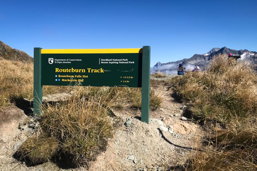 A sign post for the Routeburn Track New Zealand, with mountains and a couple of people in the background.