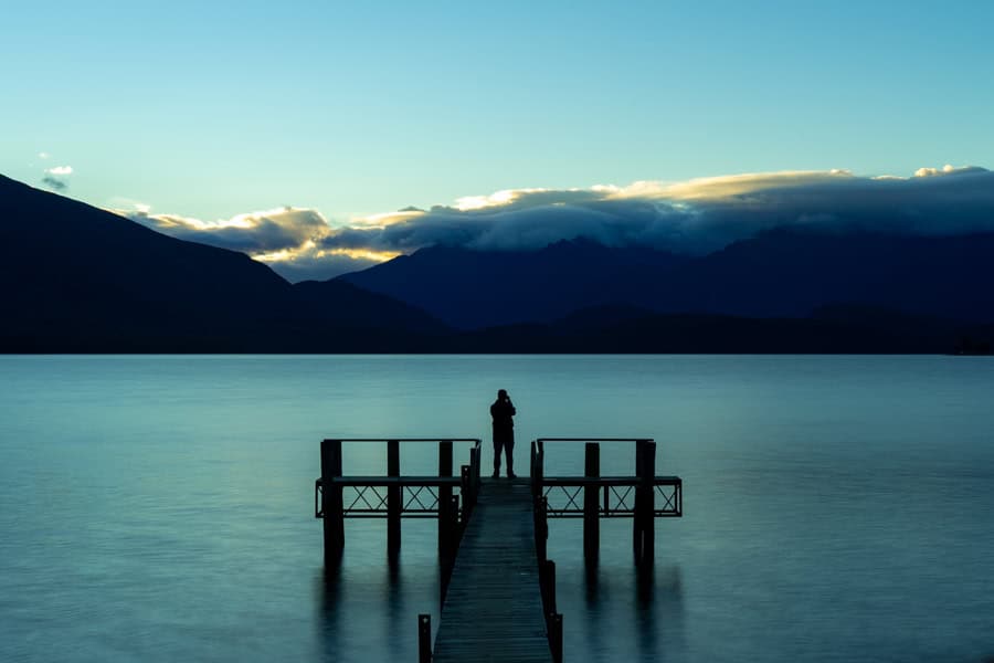 A person stands on a pier above a lake with mountains in the distance at dawn.